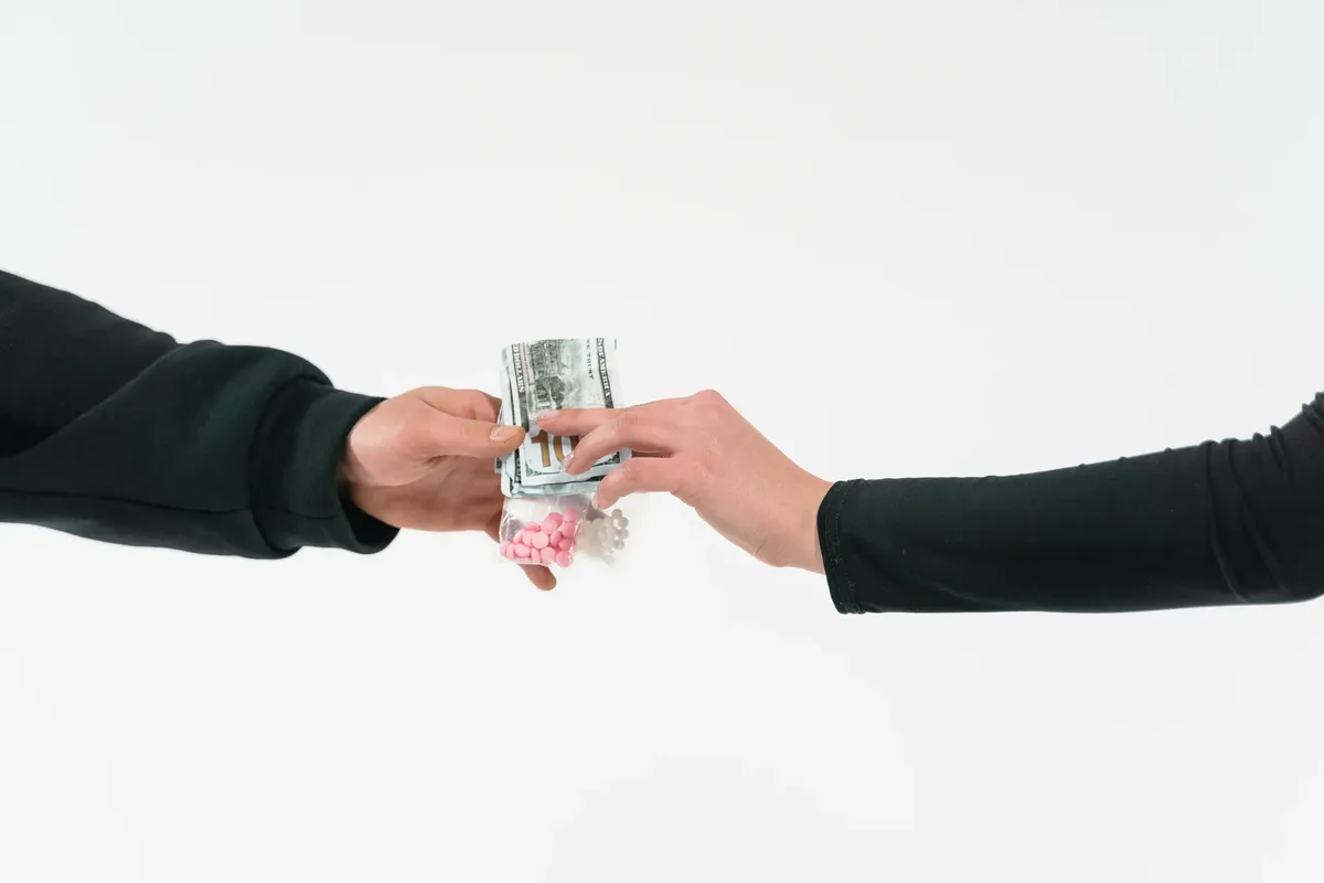 Two hands exchange pills and cash, symbolizing a transaction against a white backdrop.