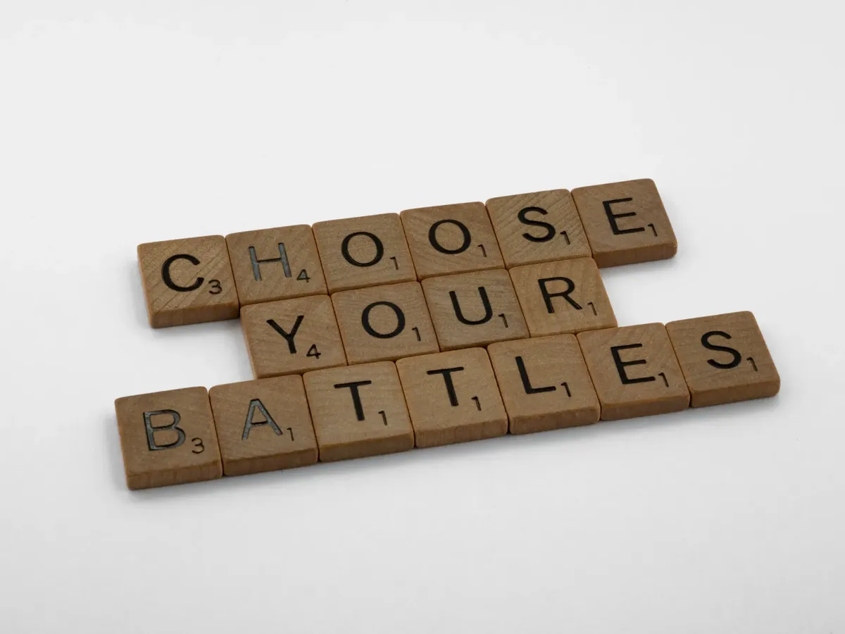 Wooden scrabble tiles spelling 'Choose Your Battles' on a white background.